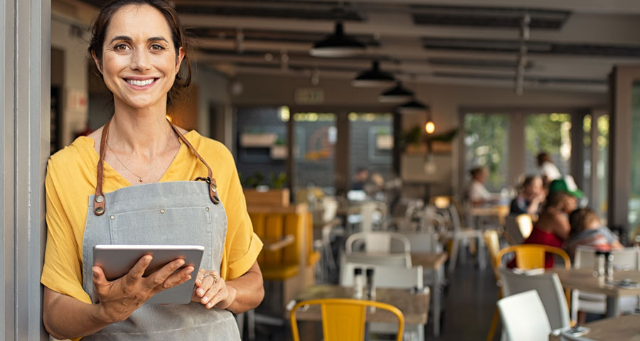 Porträt einer glücklichen Frau, die vor der Tür ihres Ladens steht. Fröhliche, reife Kellnerin wartet auf die Kunden in der Cafeteria. Erfolgreicher Kleinunternehmer mit beiläufiger Grauschürze am Eingang